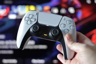 A person holding PlayStation 5 dualsense controller with game running on monitor in the background.