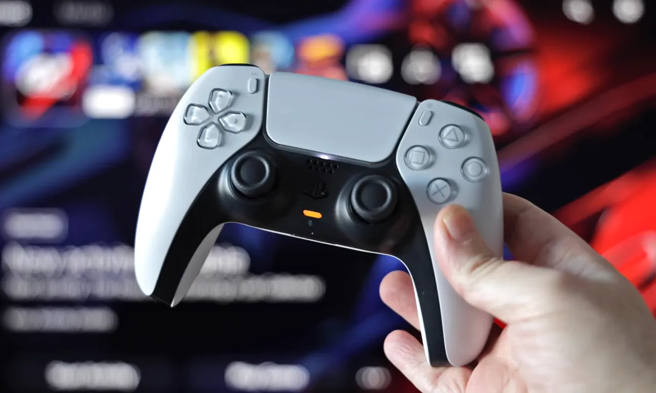 A person holding PlayStation 5 dualsense controller with game running on monitor in the background.